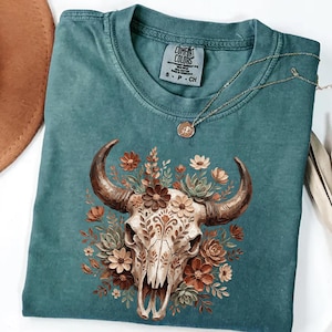 May include: Teal Comfort Colors t-shirt featuring a detailed graphic of a cow skull adorned with floral elements in shades of brown, orange, and cream. The shirt has a crew neck and short sleeves. A gold necklace with a pendant is draped across the shirt.
