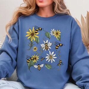 May include: A blue sweatshirt featuring a floral and bee design. The graphic includes yellow sunflowers, white daisies, green leaves, and black and yellow bees. The sweatshirt has long sleeves and a crew neck.