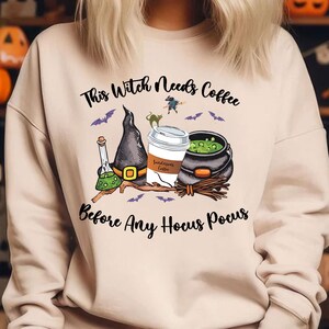 May include: Beige sweatshirt with a Halloween-themed graphic. The graphic includes a witch's hat, a coffee cup, a cauldron, and the text "This Witch Needs Coffee Before Any Hocus Pocus". Bats and a witch on a broom are also featured.