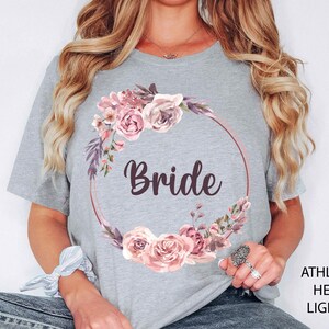 May include: Light grey t-shirt with a floral wreath design and the word "Bride" in a script font. The wreath features pink roses and purple accents. The shirt is made of a soft, heathered fabric.
