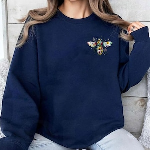 May include: A navy blue sweatshirt featuring a colorful embroidered bee design on the upper left chest. The bee is composed of various flowers and leaves. The sweatshirt has long sleeves and a crew neck.