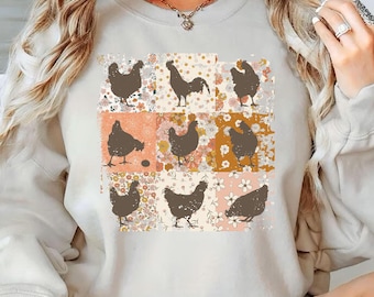 May include: Cream-colored sweatshirt featuring a grid of nine squares with chicken silhouettes. The squares have floral and solid color backgrounds in shades of orange, pink, and cream. The sweatshirt has long sleeves and a crew neck.