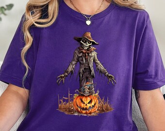 May include: A purple t-shirt featuring a Halloween-themed graphic. The design includes a scarecrow figure emerging from a carved pumpkin. The scarecrow wears a hat and tattered clothing. The pumpkin has a carved face.