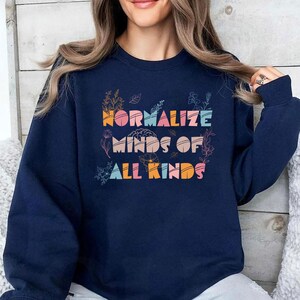 May include: Navy blue sweatshirt with the text "NORMALIZE MINDS OF ALL KINDS" in colorful, stylized letters. Floral illustrations surround the text. The sweatshirt is a long-sleeved, crew neck style.