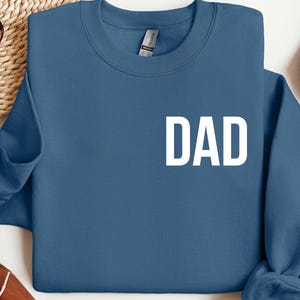 May include: A blue sweatshirt with the word "DAD" printed in large, white capital letters. The sweatshirt has a crew neck and long sleeves. The image is a close-up shot of the sweatshirt.