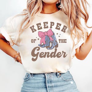 May include: Cream-colored t-shirt with the text "Keeper of the Gender" in brown lettering. The design includes a pink bow and blue cowboy boots with a pink polka dot pattern. The shirt is a casual style.