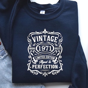 May include: Navy blue sweatshirt with a white graphic that reads "VINTAGE 1971 LIMITED EDITION Aged to PERFECTION". The design is in a decorative, ornate style. The sweatshirt is a crew neck style.