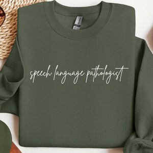 May include: An olive green sweatshirt with the words "speech language pathologist" in white script. The sweatshirt has a crew neck and long sleeves. The Gildan brand tag is visible on the inside of the collar.