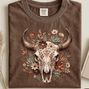 Puede incluir: Camiseta color espresso con una ilustraci&oacute;n detallada de una calavera de vaca adornada con elementos florales en tonos marrones, crema y verdes. La camiseta tiene un cuello redondo cl&aacute;sico y una etiqueta Comfort Colors.