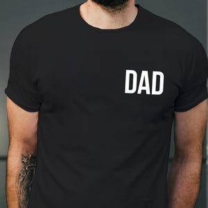 May include: Black t-shirt with the white word "DAD" printed on the chest. The shirt has short sleeves and a crew neck. The image shows the upper body of a person wearing the shirt.