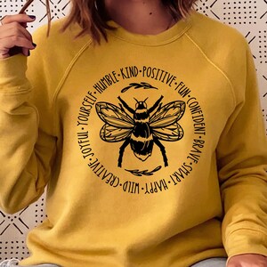 May include: Mustard yellow sweatshirt with a black bee graphic in a circular design. The design includes the words: "Joyful, Yourself, Humble, Kind, Positive, Fun, Confident, Brave, Smart, Happy, Wild, Creative."