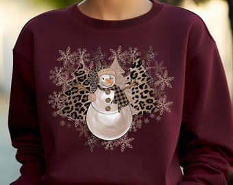 May include: Burgundy sweatshirt featuring a snowman design. The snowman has a carrot nose, a plaid scarf, and a hat. The design includes leopard print Christmas trees and snowflakes. A festive winter-themed apparel item.