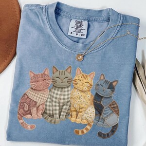 May include: A light blue Comfort Colors t-shirt featuring four smiling cat illustrations. Each cat has a unique patchwork design in shades of pink, gray, yellow, and blue. A gold necklace with a pendant rests on the shirt.