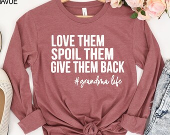 May include: A heather mauve long-sleeve shirt featuring white text that reads LOVE THEM SPOIL THEM GIVE THEM BACK #grandma life. The shirt is displayed on a wooden hanger with a round neckline and long sleeves, showcasing a casual design.