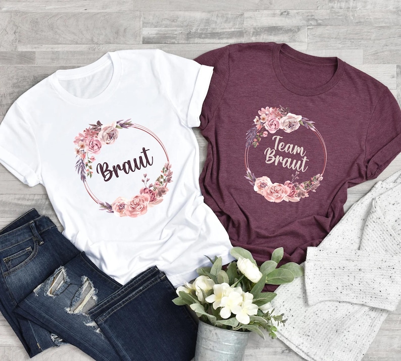 Puede incluir: Dos camisetas, una blanca y otra burdeos, con dise&ntilde;os de coronas florales. La camiseta blanca dice "Braut" en cursiva, y la camiseta burdeos dice "Team Braut". Ambas camisetas son de manga corta y tienen cuello redondo. Tambi&eacute;n se ven unos vaqueros azul oscuro y un peque&ntilde;o ramo de flores.