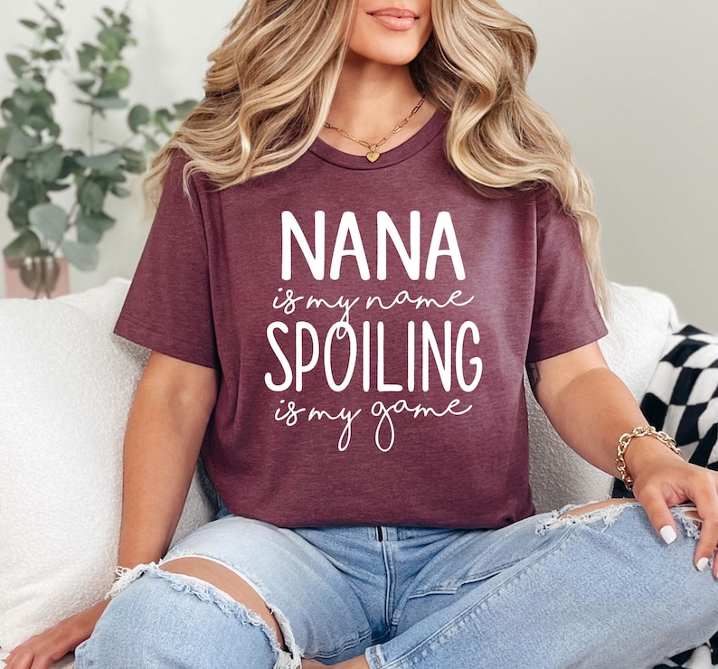May include: Burgundy t-shirt with the text "NANA is my name, SPOILING is my game" in white. The shirt has a crew neck and short sleeves. The person is wearing blue jeans.