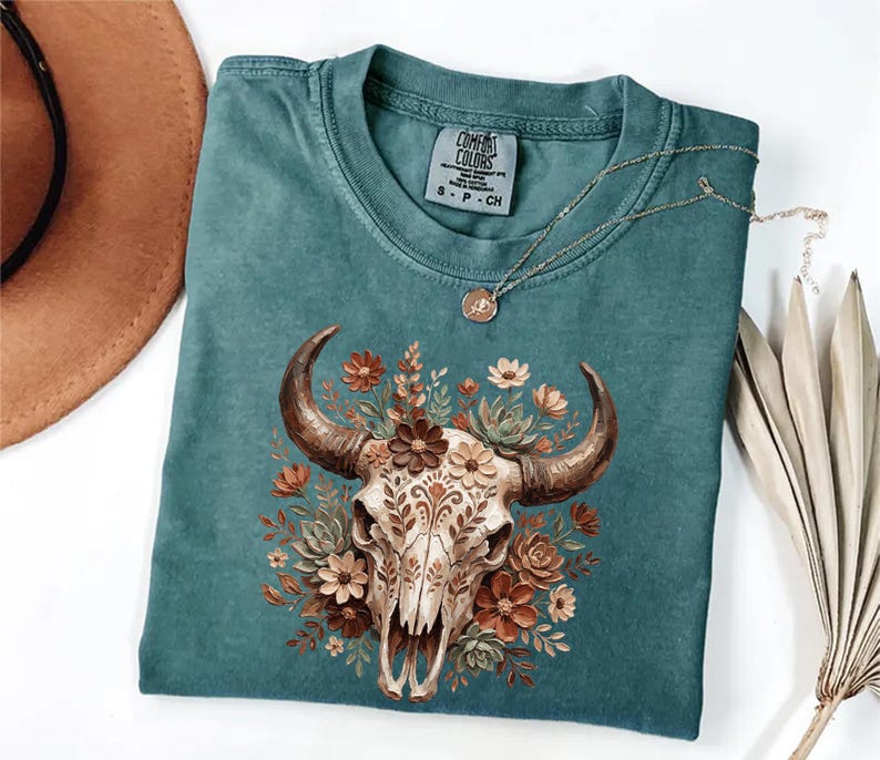 May include: Teal Comfort Colors t-shirt featuring a detailed graphic of a cow skull adorned with floral elements in shades of brown, orange, and cream. The shirt has a crew neck and short sleeves. A gold necklace with a pendant is draped across the shirt.
