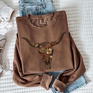 May include: A brown long-sleeve crewneck sweatshirt featuring a graphic of a cow skull filled with various orange, yellow, and brown flowers. The inner neck label displays the text Comfort Colors and the size L G.