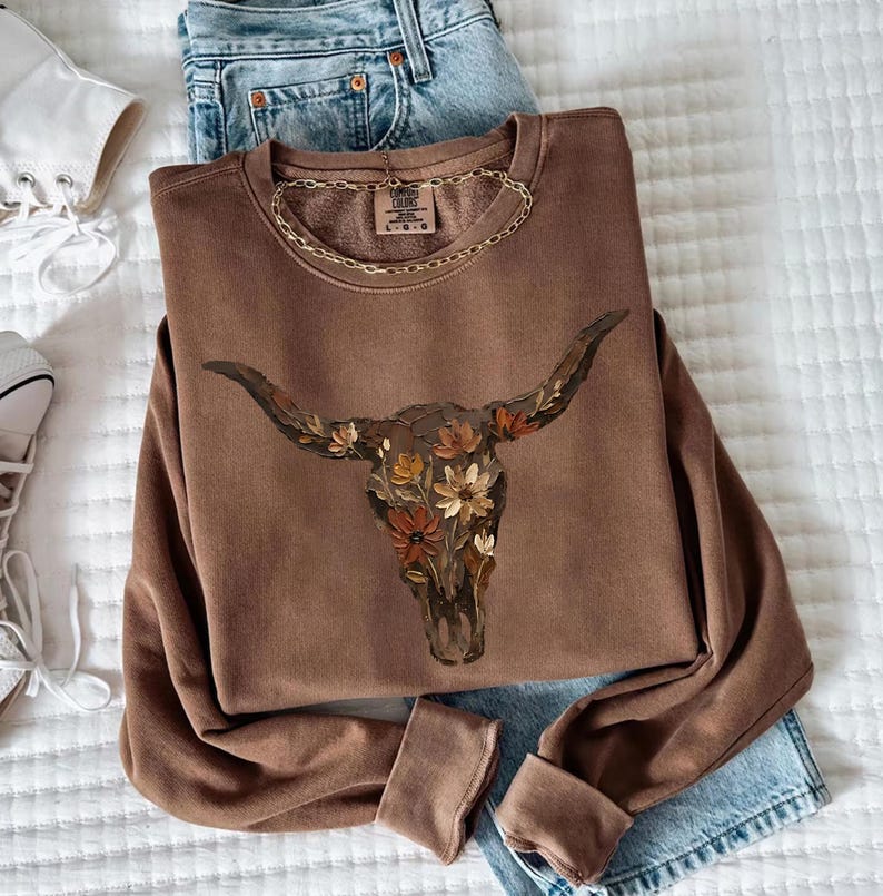 May include: A brown long-sleeve crewneck sweatshirt featuring a graphic of a cow skull filled with various orange, yellow, and brown flowers. The inner neck label displays the text Comfort Colors and the size L G.