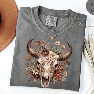 Puede incluir: Una camiseta gris Comfort Colors con un dise&ntilde;o de calavera de toro adornada con flores marrones, naranjas y crema. La camiseta tiene cuello redondo y mangas cortas. Un collar plateado con un colgante est&aacute; drapeado sobre la camiseta.