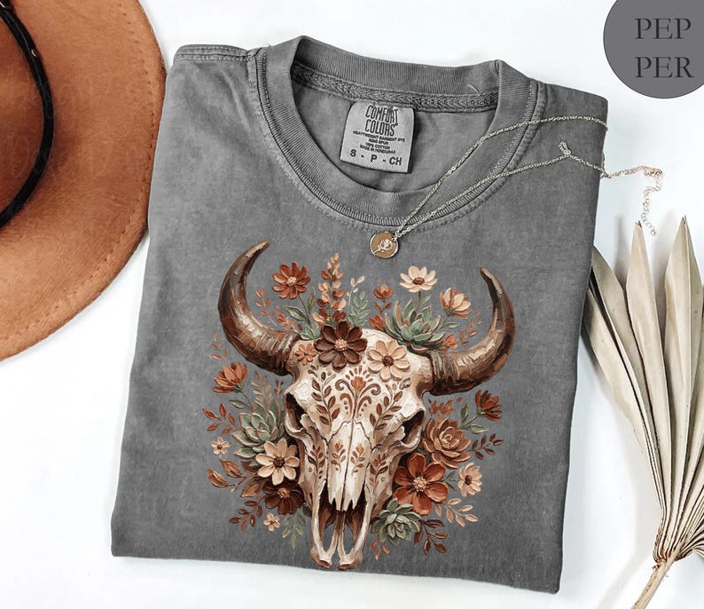 Puede incluir: Una camiseta gris Comfort Colors con un dise&ntilde;o de calavera de toro adornada con flores marrones, naranjas y crema. La camiseta tiene cuello redondo y mangas cortas. Un collar plateado con un colgante est&aacute; drapeado sobre la camiseta.