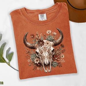 Puede incluir: Una camiseta naranja Comfort Colors con una ilustraci&oacute;n detallada de una calavera de toro adornada con flores y suculentas. La calavera es de color crema con patrones intrincados, y los cuernos son de color marr&oacute;n oscuro. La camiseta tiene un cuello redondo cl&aacute;sico.
