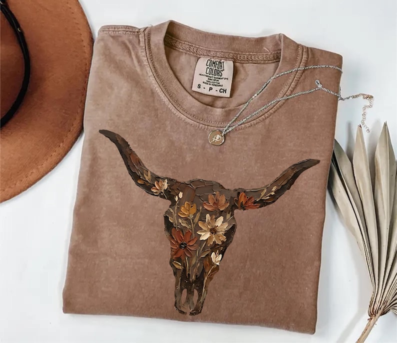 May include: A brown short-sleeve t-shirt featuring a graphic of a cow skull adorned with painted flowers in shades of orange, yellow, and brown. The shirt has a Comfort Colors brand label at the neck with size S, and a silver necklace rests on top.