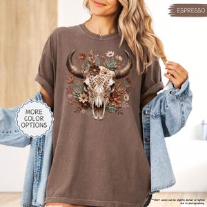 Puede incluir: Camiseta color espresso con un dise&ntilde;o gr&aacute;fico detallado de una calavera de toro adornada con flores en tonos marrones y verdes. La calavera tiene patrones intrincados. La camiseta tiene mangas enrolladas y un corte holgado. El texto "MORE COLOR OPTIONS" es visible.