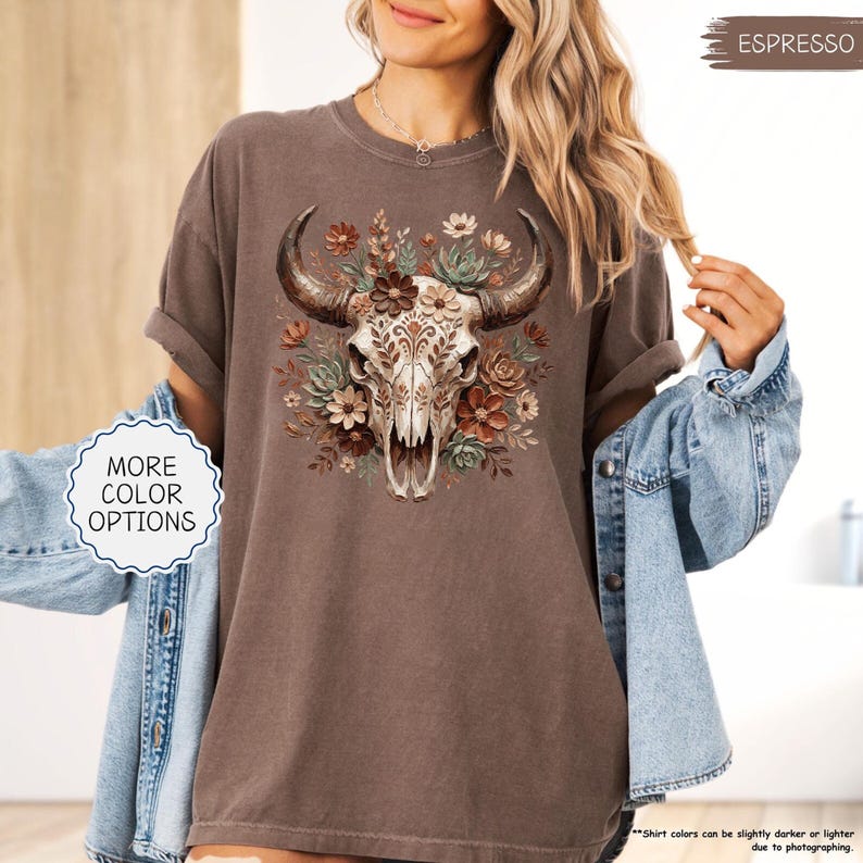 Puede incluir: Camiseta color espresso con un dise&ntilde;o gr&aacute;fico detallado de una calavera de toro adornada con flores en tonos marrones y verdes. La calavera tiene patrones intrincados. La camiseta tiene mangas enrolladas y un corte holgado. El texto "MORE COLOR OPTIONS" es visible.