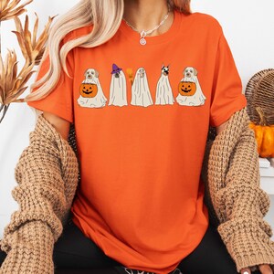 May include: Orange t-shirt featuring a Halloween-themed design. The graphic depicts five dogs dressed as ghosts, each with unique accessories, including a witch hat and a carved pumpkin. The dogs are in a row, creating a festive and playful image.