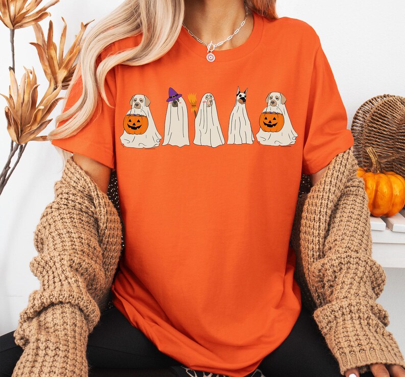 May include: Orange t-shirt featuring a Halloween-themed design. The graphic depicts five dogs dressed as ghosts, each with unique accessories, including a witch hat and a carved pumpkin. The dogs are in a row, creating a festive and playful image.