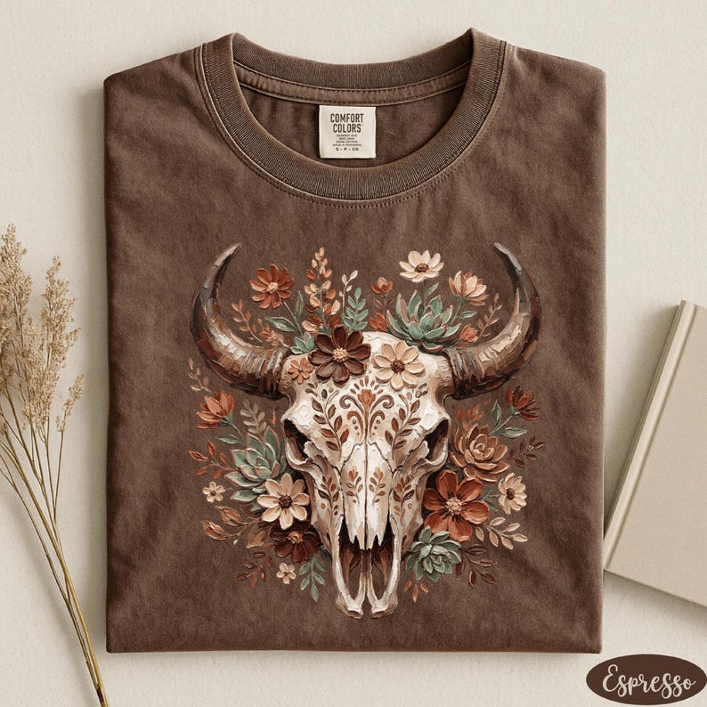 Puede incluir: Camiseta color espresso con una ilustraci&oacute;n detallada de una calavera de vaca adornada con elementos florales en tonos marrones, crema y verdes. La camiseta tiene un cuello redondo cl&aacute;sico y una etiqueta Comfort Colors.