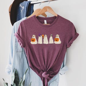 May include: A maroon t-shirt featuring a Halloween-themed graphic. The design includes cartoon dogs dressed as ghosts, a witch, and holding jack-o'-lanterns. The shirt is hanging on a wooden hanger with other clothing items in the background.