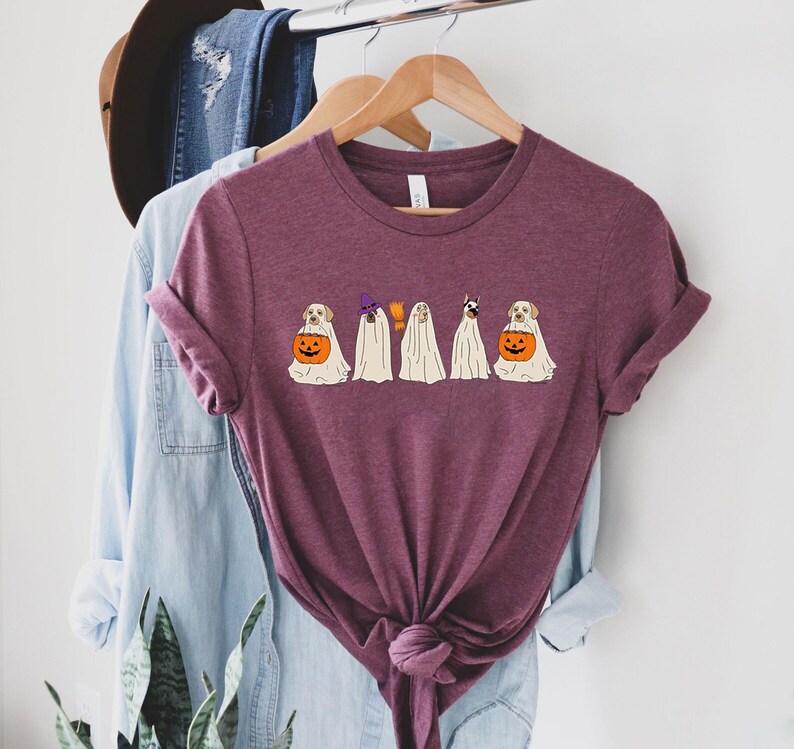 May include: A maroon t-shirt featuring a Halloween-themed graphic. The design includes cartoon dogs dressed as ghosts, a witch, and holding jack-o'-lanterns. The shirt is hanging on a wooden hanger with other clothing items in the background.