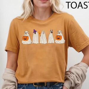 May include: Mustard-colored t-shirt featuring a Halloween-themed graphic. The graphic depicts five dogs dressed as ghosts, with one holding a jack-o'-lantern. The word "TOAST" is printed in black at the top right corner.
