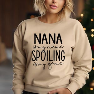 May include: A beige sweatshirt with the text "NANA is my name, SPOILING is my game" in black script. The sweatshirt has a crew neck and long sleeves. The background is blurred, with hints of a Christmas tree.