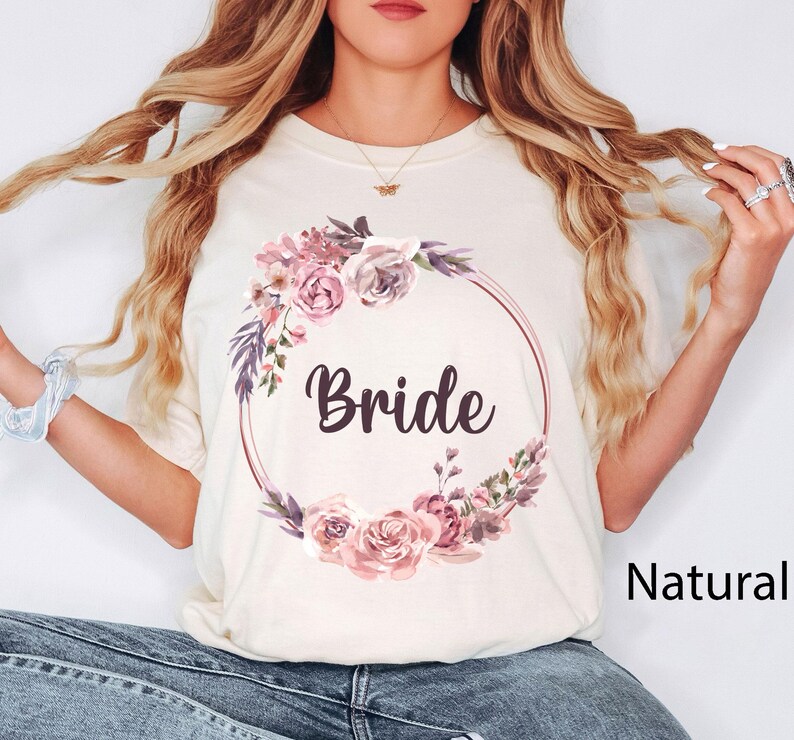 Puede incluir: Camiseta color crema con la palabra "Bride" en fuente oscura, rodeada por una corona floral en tonos rosas y morados. La camiseta tiene cuello redondo y mangas cortas, perfecta para un evento nupcial.