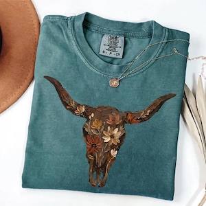 May include: A teal short-sleeved t-shirt featuring a graphic of a brown cow skull adorned with painted flowers in shades of orange, yellow, and white. The shirt has a crew neckline and a Comfort Colors brand label printed on the inside neck area.
