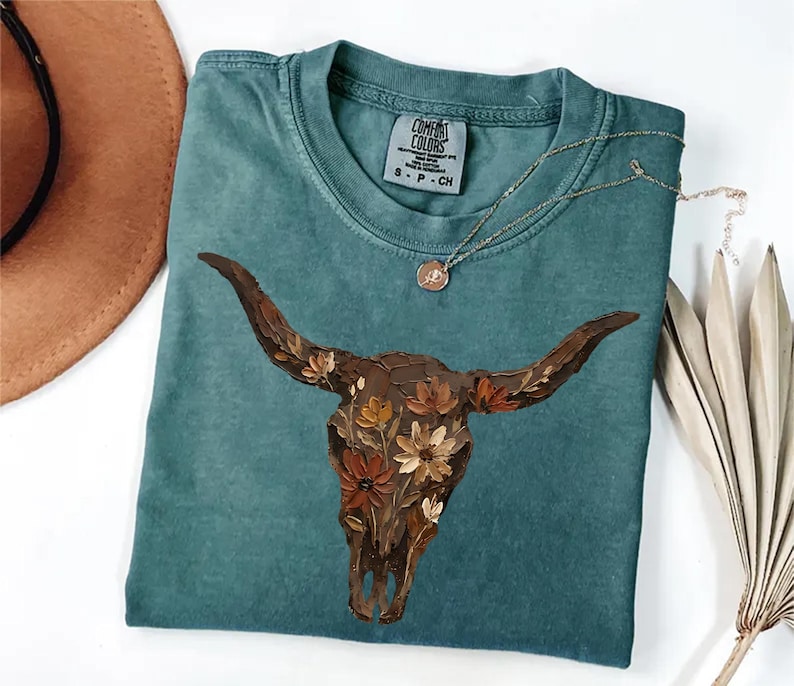 May include: A teal short-sleeved t-shirt featuring a graphic of a brown cow skull adorned with painted flowers in shades of orange, yellow, and white. The shirt has a crew neckline and a Comfort Colors brand label printed on the inside neck area.