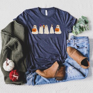 May include: Navy blue t-shirt featuring a Halloween design with dogs dressed as ghosts, holding pumpkin baskets. The shirt is styled with blue jeans, brown boots, and a green sweater. A white and red pumpkin are also in the image.