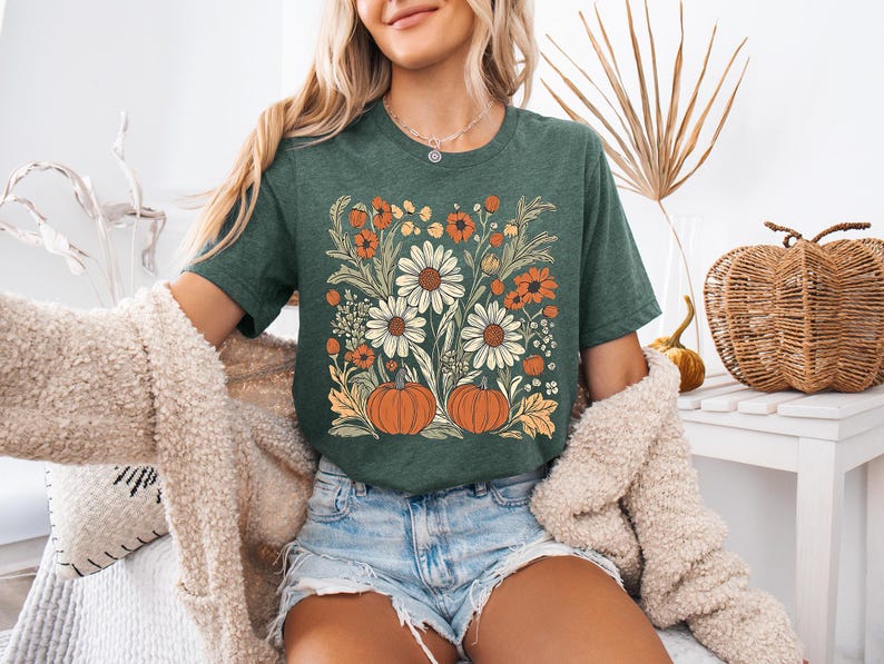 May include: Green t-shirt featuring a floral design with white daisies, orange flowers, and two orange pumpkins. The shirt is a short-sleeved crew neck. The design has a vintage, fall-themed aesthetic.