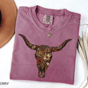May include: A berry-coloured t-shirt featuring a graphic of a brown longhorn skull adorned with orange, yellow, and brown flowers. The shirt has a crew neckline and a Comfort Colors brand label on the inside collar with the size S P CH printed.