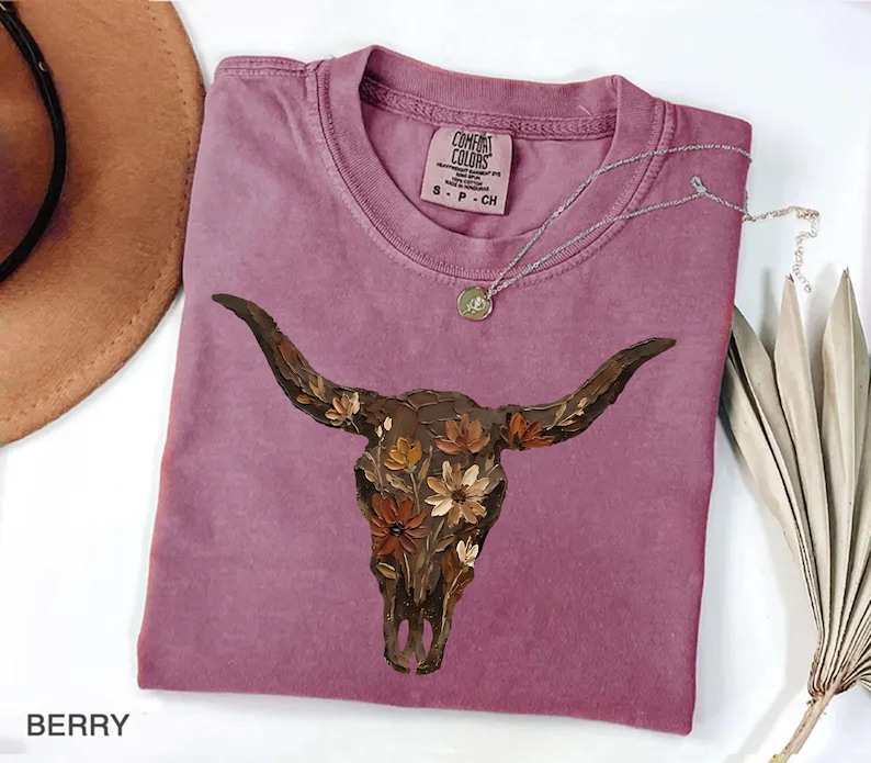 May include: A berry-coloured t-shirt featuring a graphic of a brown longhorn skull adorned with orange, yellow, and brown flowers. The shirt has a crew neckline and a Comfort Colors brand label on the inside collar with the size S P CH printed.