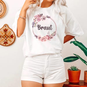 Puede incluir: Camiseta blanca con un dise&ntilde;o de corona floral y la palabra "Braut" en escritura oscura. La corona presenta flores rosas y moradas y hojas verdes. La camiseta se combina con pantalones cortos blancos.