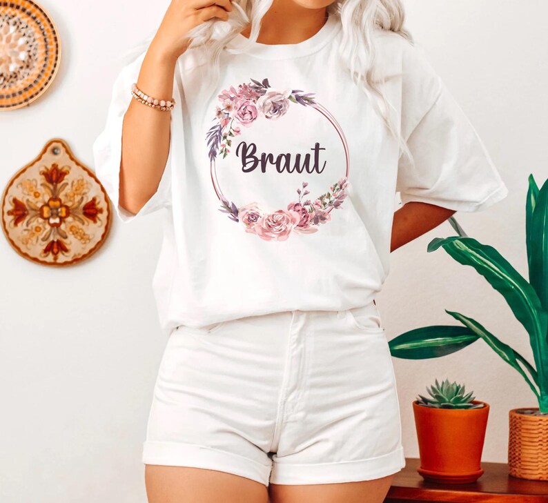 Puede incluir: Camiseta blanca con un dise&ntilde;o de corona floral y la palabra "Braut" en escritura oscura. La corona presenta flores rosas y moradas y hojas verdes. La camiseta se combina con pantalones cortos blancos.