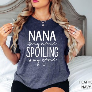 May include: Heather navy t-shirt with the text "NANA is my name, SPOILING is my game" in white script. The shirt is a dark blue-grey color and has a crew neck. The words "Heather Navy" are printed in the bottom right corner.
