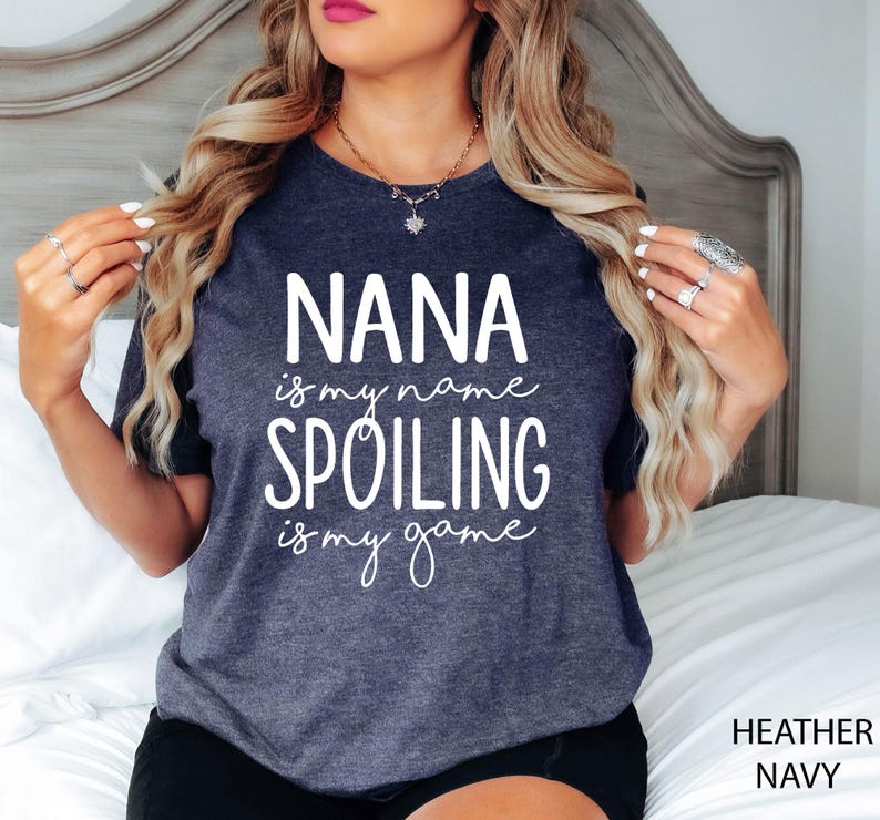 May include: Heather navy t-shirt with the text "NANA is my name, SPOILING is my game" in white script. The shirt is a dark blue-grey color and has a crew neck. The words "Heather Navy" are printed in the bottom right corner.