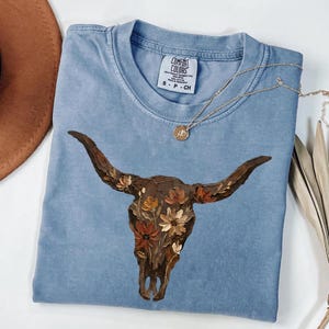 May include: A blue short-sleeved t-shirt featuring a graphic of a brown longhorn skull adorned with painted flowers in shades of orange, yellow, and cream. The shirt has a crew neckline and a Comfort Colors brand label printed on the inside collar.