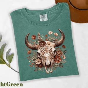 Puede incluir: Camiseta verde claro con una ilustraci&oacute;n detallada de una calavera de toro adornada con flores en tonos marr&oacute;n, naranja y crema. La calavera tiene patrones intrincados. Un sombrero marr&oacute;n es visible en el fondo. La camiseta est&aacute; etiquetada como "Comfort Colors".