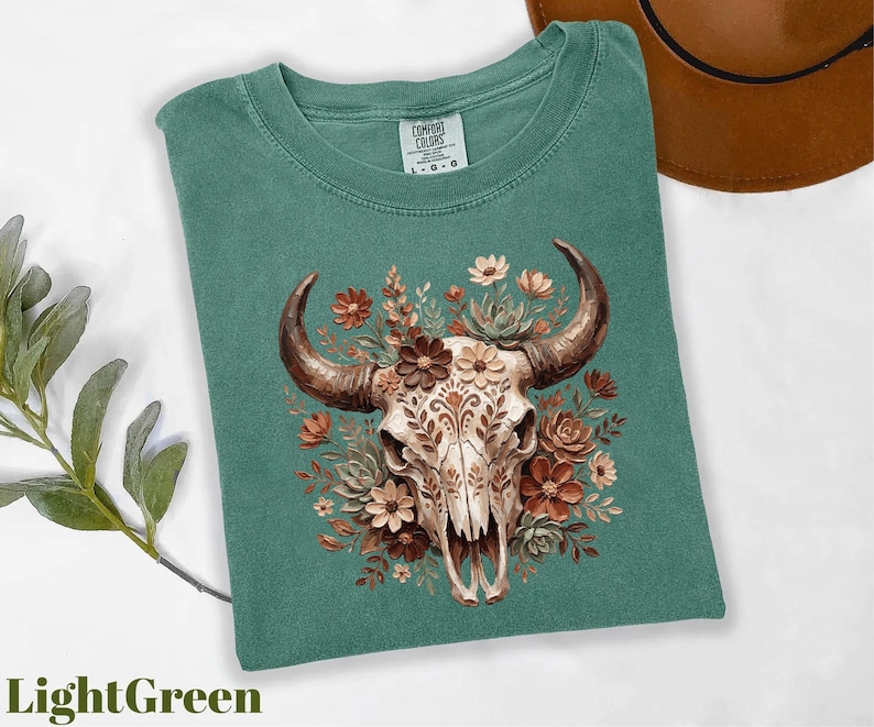Puede incluir: Camiseta verde claro con una ilustraci&oacute;n detallada de una calavera de toro adornada con flores en tonos marr&oacute;n, naranja y crema. La calavera tiene patrones intrincados. Un sombrero marr&oacute;n es visible en el fondo. La camiseta est&aacute; etiquetada como "Comfort Colors".